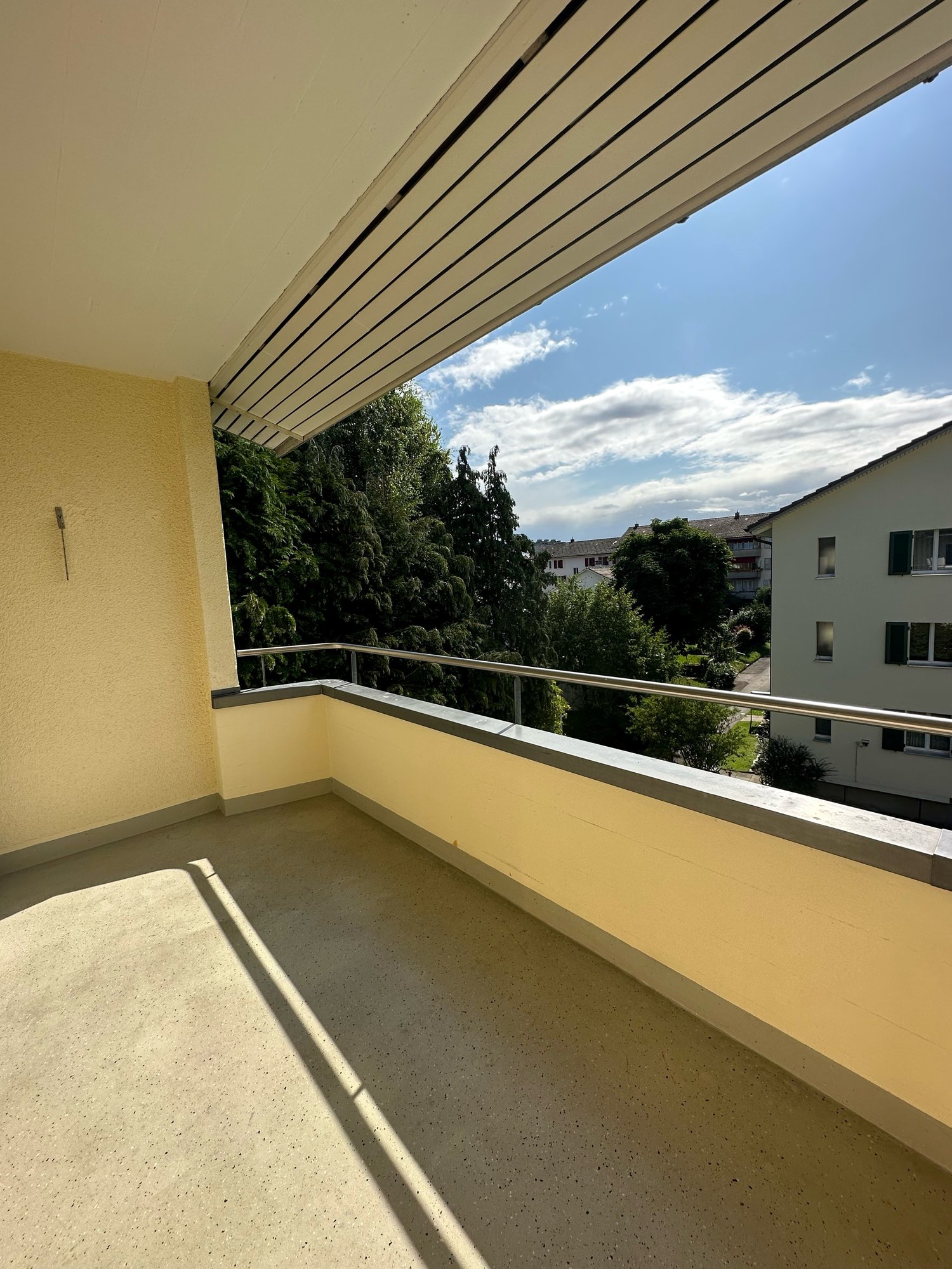 Large balcony with yellow walls, metal railings, concrete floor, and a view of trees and buildings in the distance
