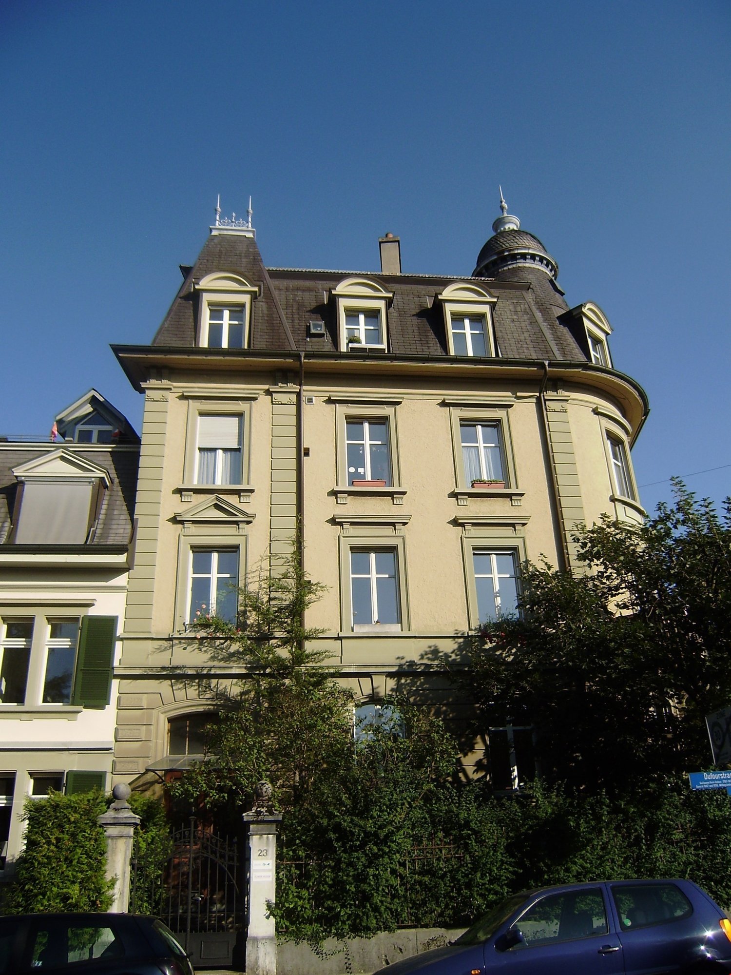 3 story building, beige color, several windows, black rooftop, 2 cars parked