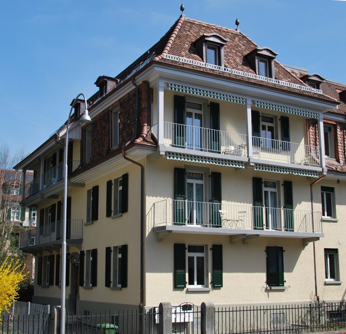 3 story building, red roof, balconies, windows with green shutters, yellow tree, street light, fence