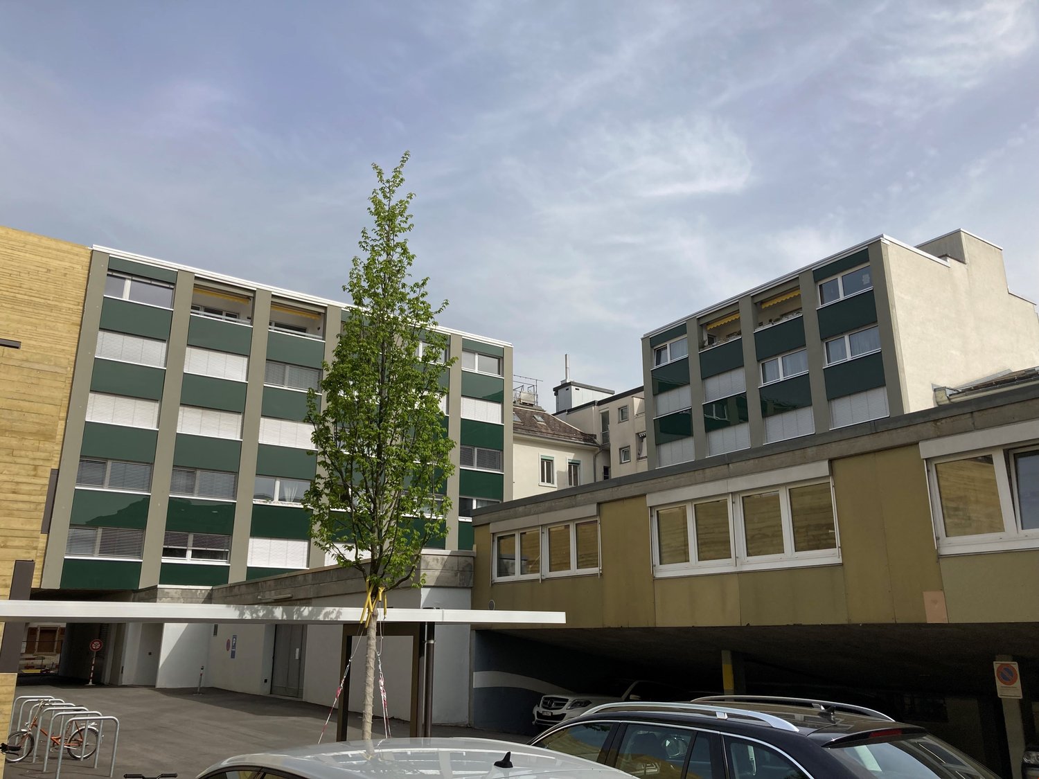 Modern multi-story residential building with large windows and green accents, cars parked in underground garage.
