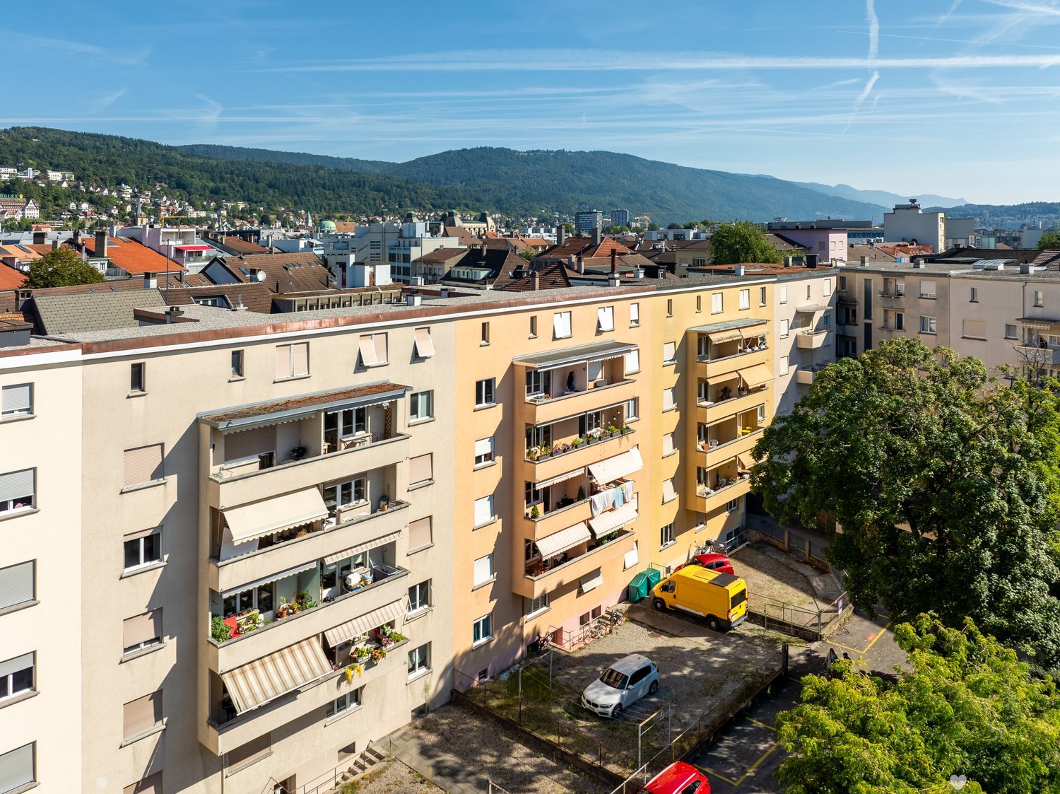 large multi-story building, multiple balconies, several parked vehicles, green space