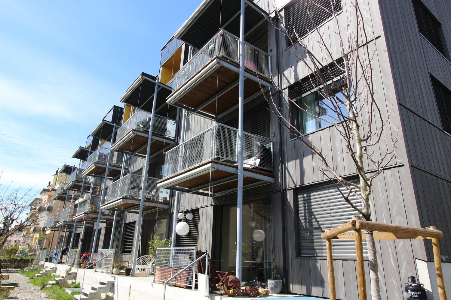 gray exterior, multiple balconies, metal railings, stairs, metal fence, potted plants, hanging lamps