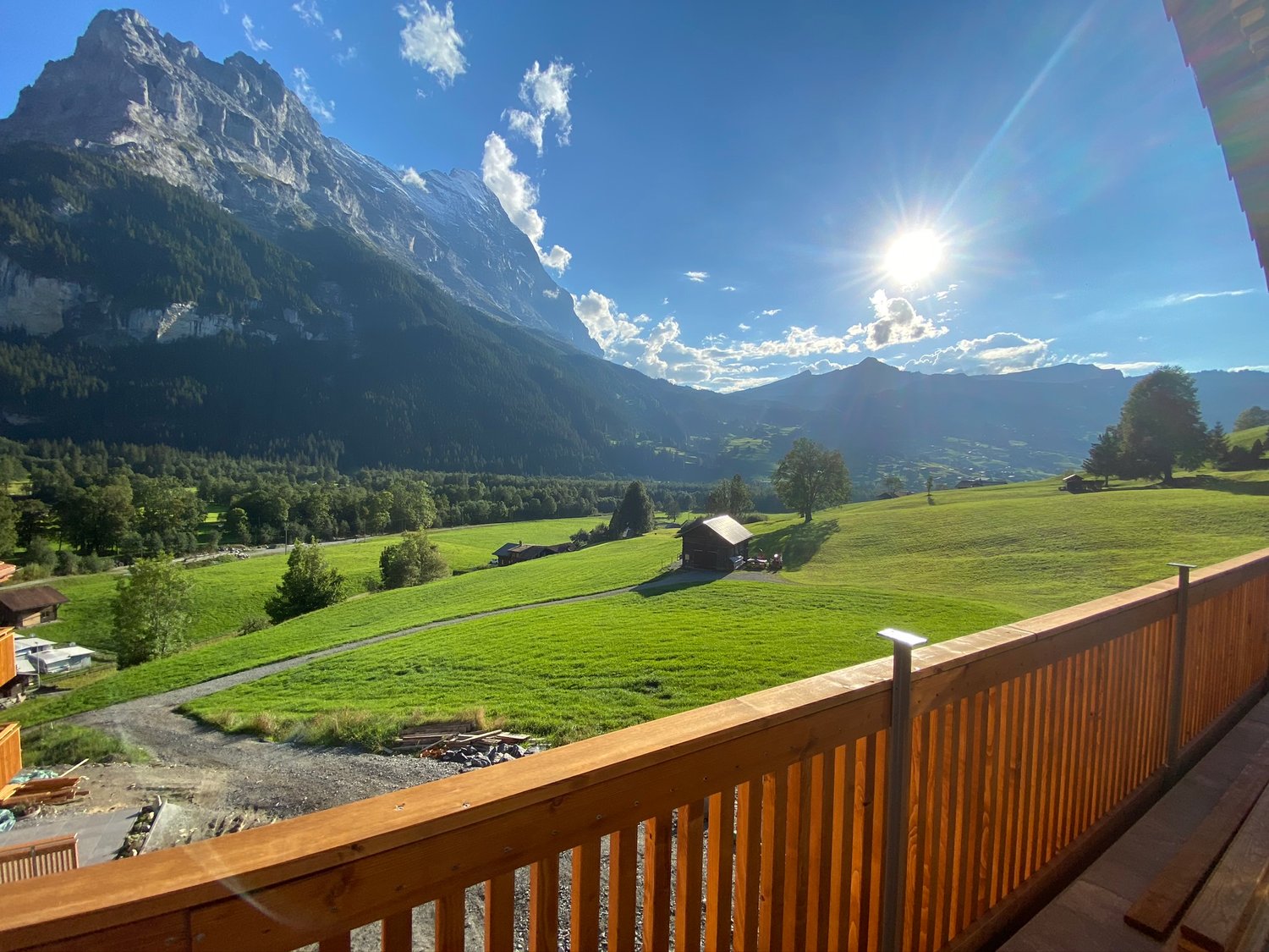 View from a wooden deck showing a vast green valley with mountains in the background, a small wooden chalet, and a clear blue sky.