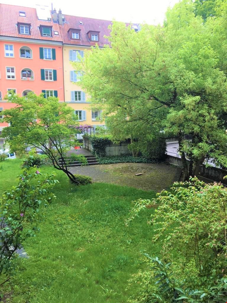 The image shows a residential building with colorful facades and a lush, green garden area in the foreground. The building has multiple floors and windows, and there appears to be a balcony or terrace visible on one of the upper floors.