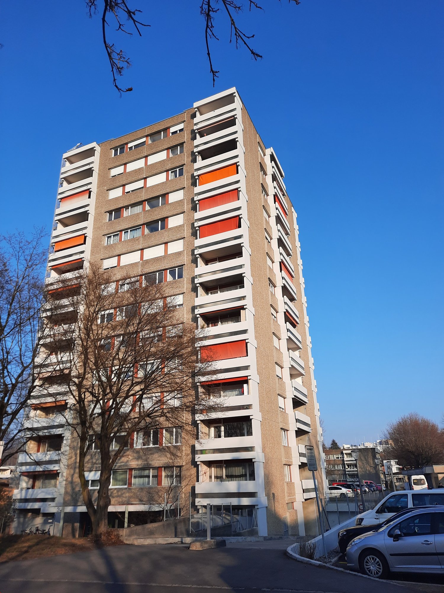 A tall residential building with multiple floors, balconies with orange shutters, and a beige and white exterior.