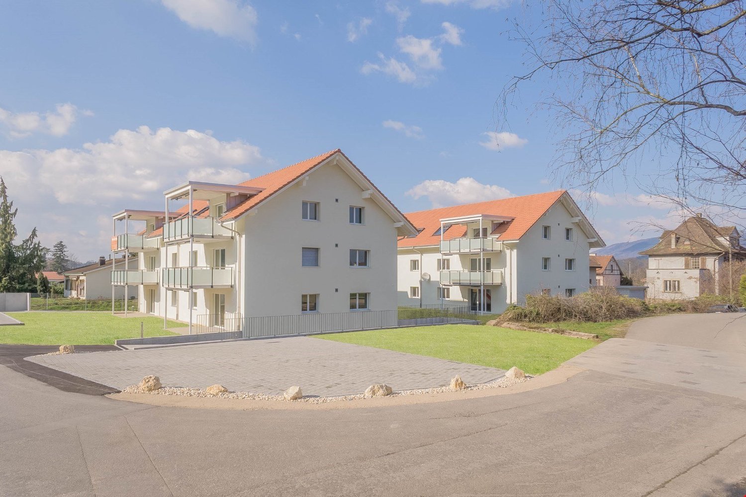 Multi-story apartment building with white exterior walls and orange tile roofs, surrounded by a grassy area with paved walkways and landscaping elements