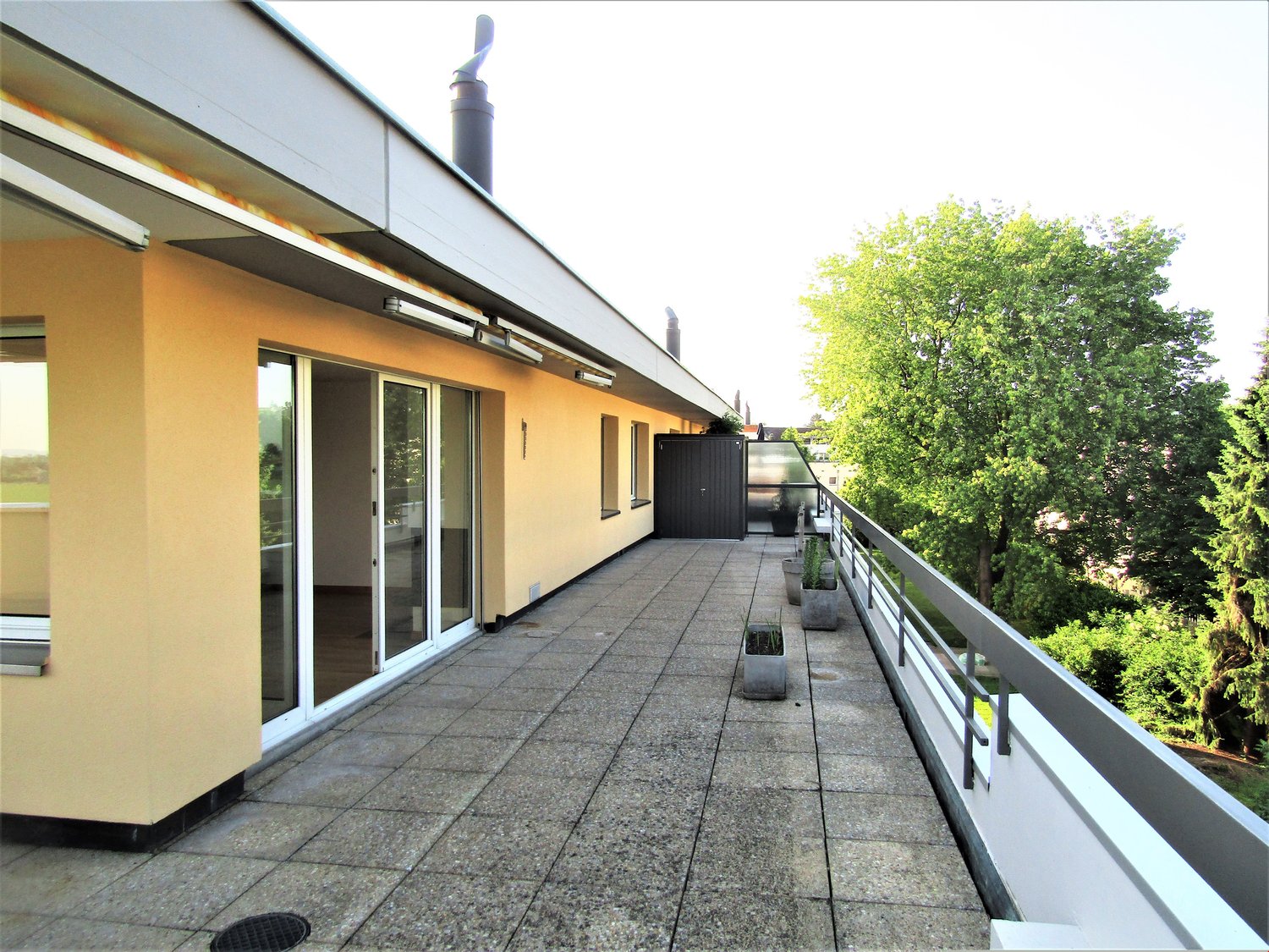 Balcony with granite tile flooring, glass sliding doors, exterior walls with beige stucco finish, trees and greenery visible in the background