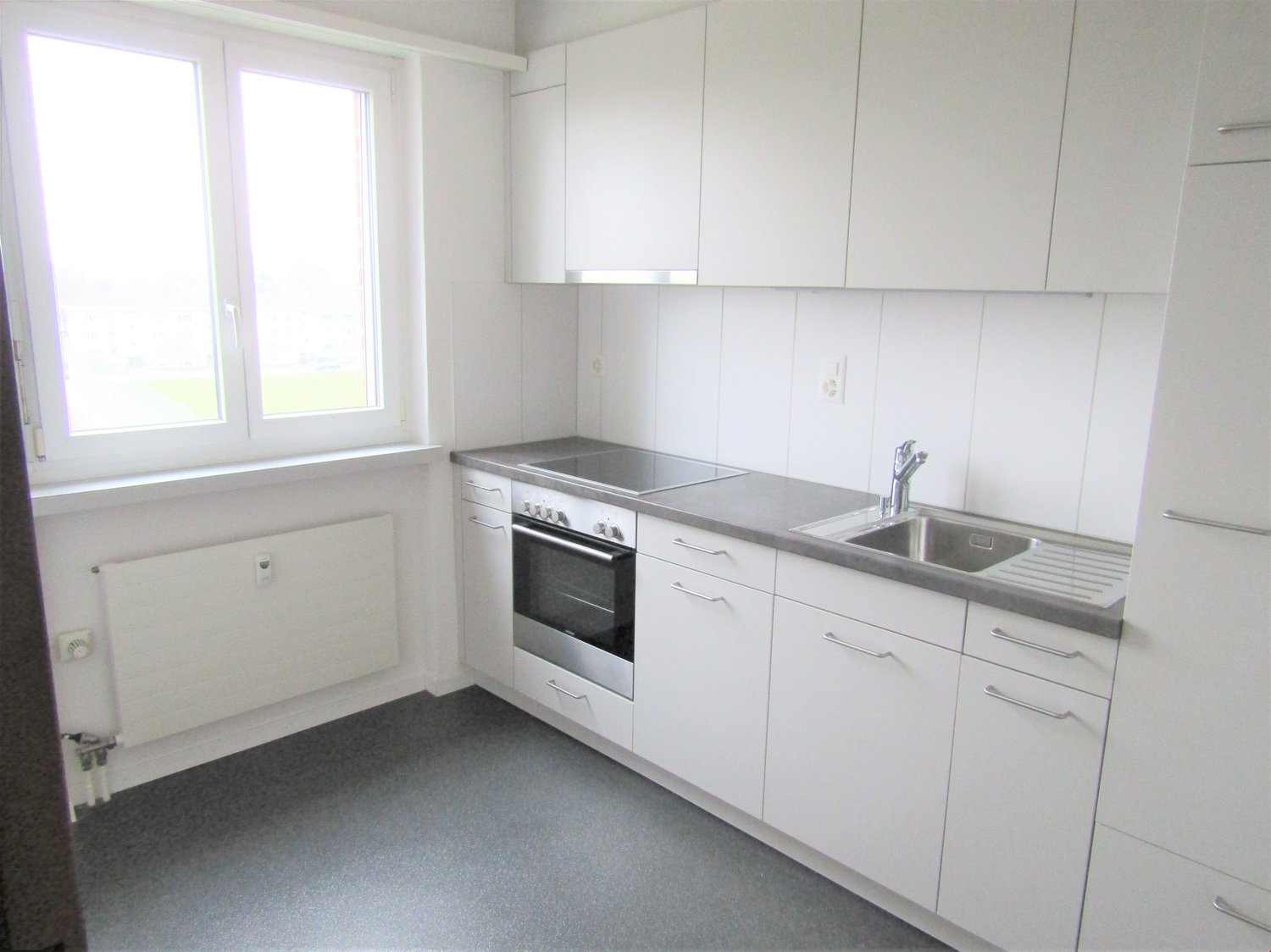 Modern kitchen with white cabinets, stainless steel countertop, built-in oven, and a sink with a faucet. It has a window with a white frame. The floor is tiled with gray tiles.