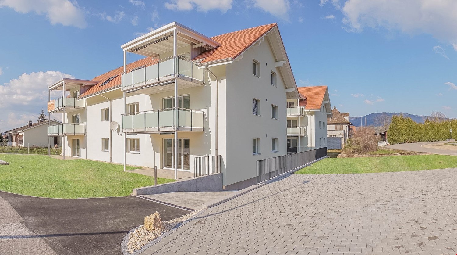 Modern 2-story white building with orange roof, multiple balconies, large windows, driveway, grassy area in front