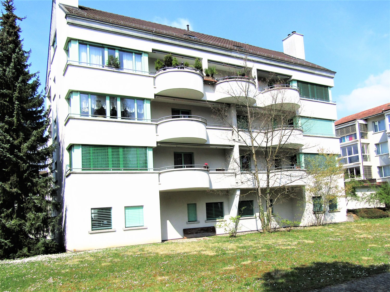 3-story apartment building with white exterior, balconies on each floor, and large windows. The building has a green roof and is surrounded by a grassy area with trees.