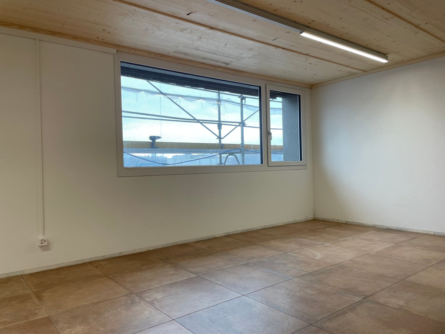 This image shows a spacious room with a high wooden ceiling and large windows that provide natural light. The room has a tiled floor and plain white walls, giving it a minimalist and clean appearance. There are no visible furniture or other features in th