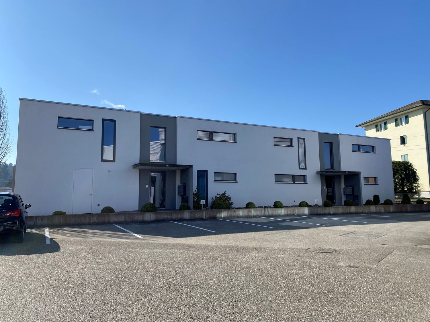 modern white building, multiple windows, parking lot in front, trees in the distance