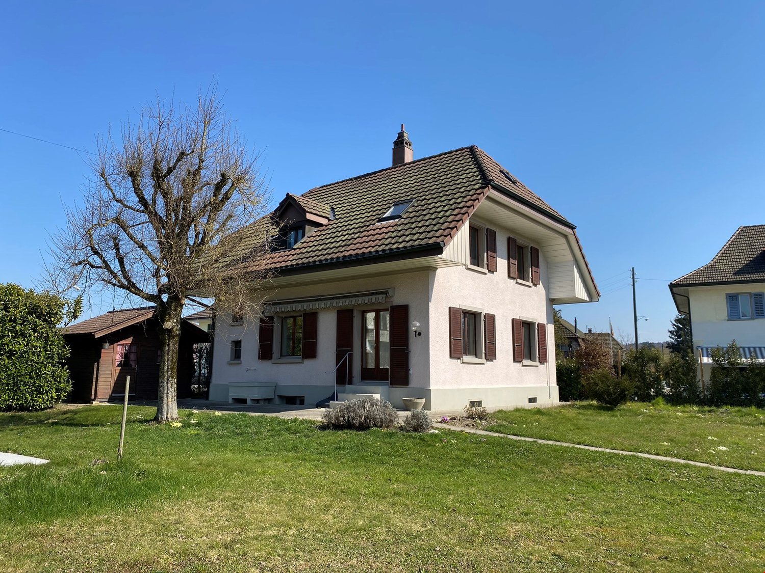 Detached house with two floors, brown roof, windows with shutters, chimney, garden with green lawn, bench at the entrance