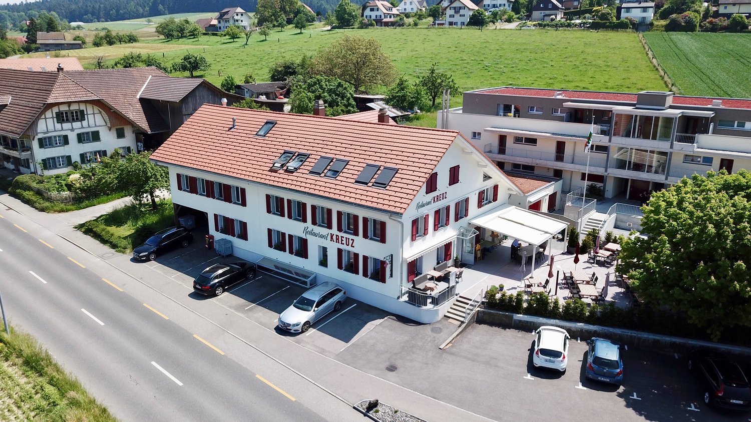 The house has a red roof, multiple windows, and a restaurant with an outdoor dining area. Cars are parked in the parking lot. In the background are trees, fields, and houses.