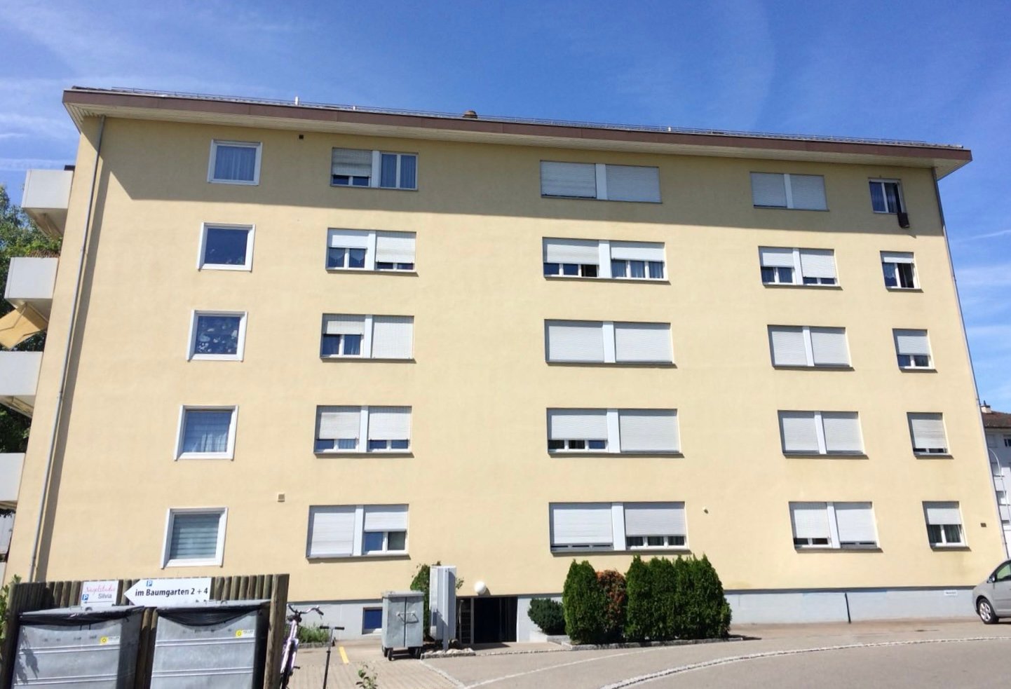 4 story building, beige exterior, white windows, small garden, trash bins, car parked