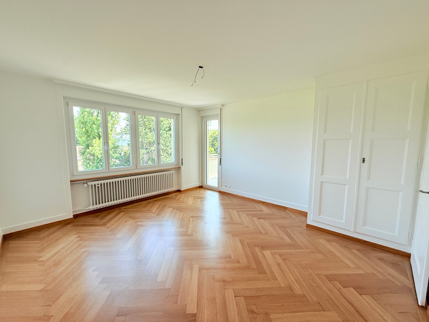 Spacious room with large windows, hardwood parquet flooring, and a radiator. The room appears to be empty, with white walls and a door leading to another area.