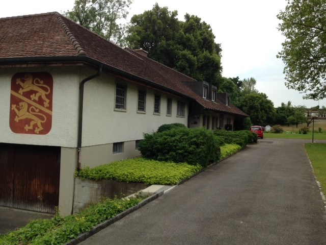 Two story building, brown roof, red emblem, white walls, driveway, wooden garage door, shrubs, trees