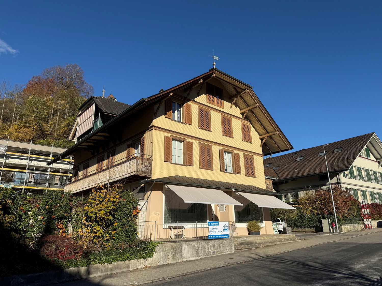 three-story building, yellow exterior, multiple windows, black roof, awnings, balcony, large bushes and plants, street view