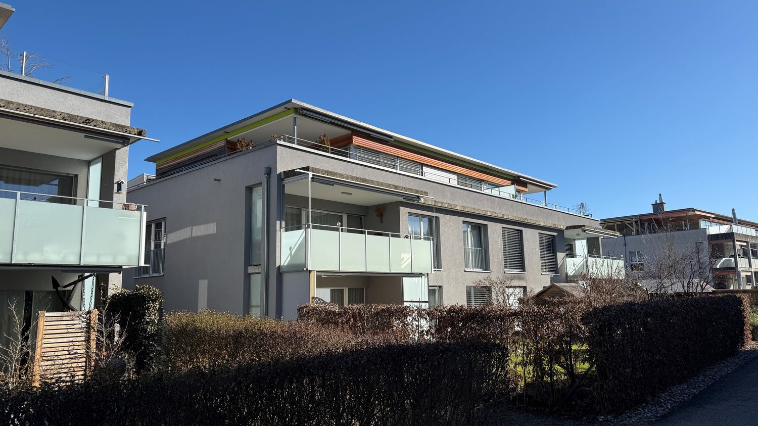 Three-story house, gray facade, glass railings on the upper floors, green roofing, covered area in the entrance, shrubs in the foreground