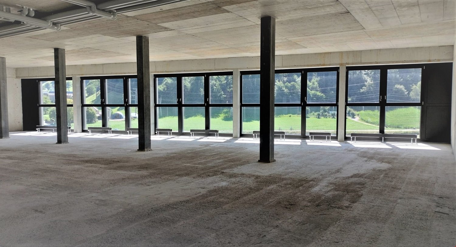 A wide empty room with many windows, two long benches, and pillars