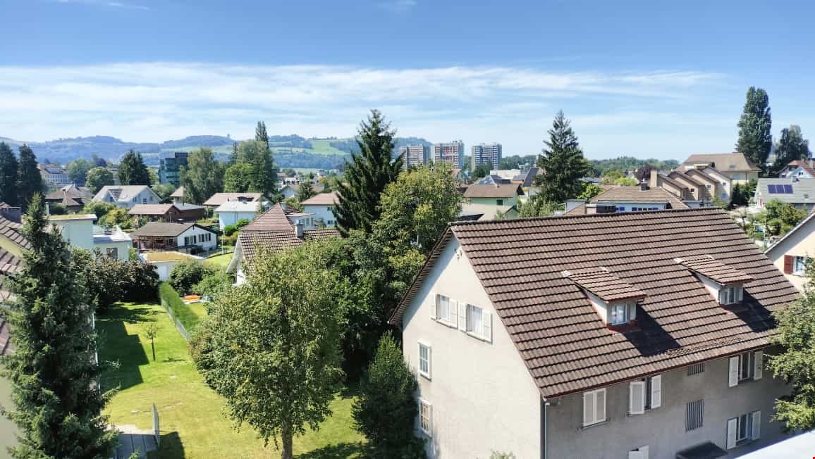 The image shows a residential neighborhood with a mix of single-family homes and multi-story apartment buildings. The area is surrounded by lush greenery, including trees and well-maintained lawns. The buildings have a mix of architectural styles, with so
