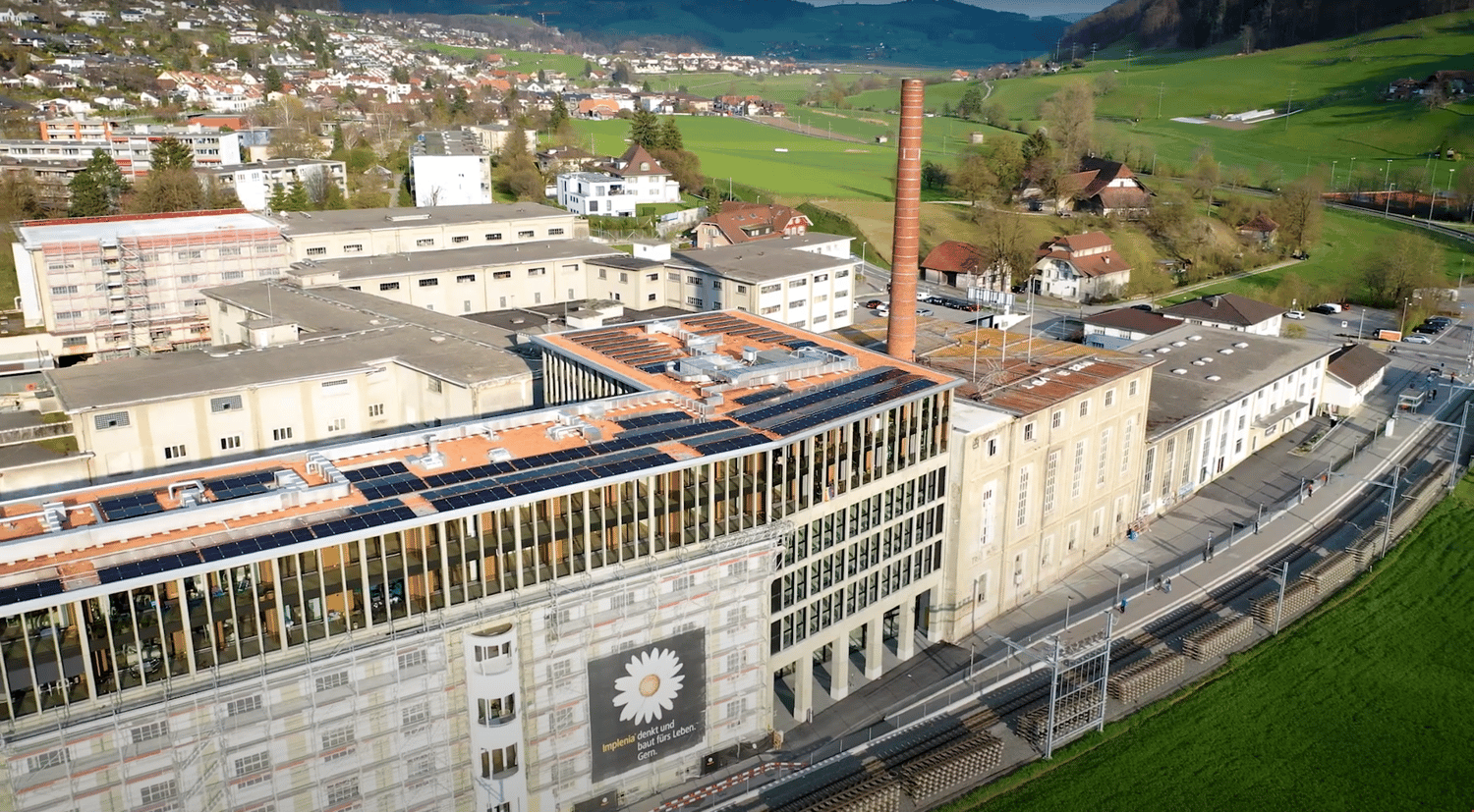 A large multi-story building complex with solar panels on the roof, surrounded by greenery and a backdrop of hills. The building features glass windows, a tall chimney, and various other architectural details. Several smaller houses and structures are vis