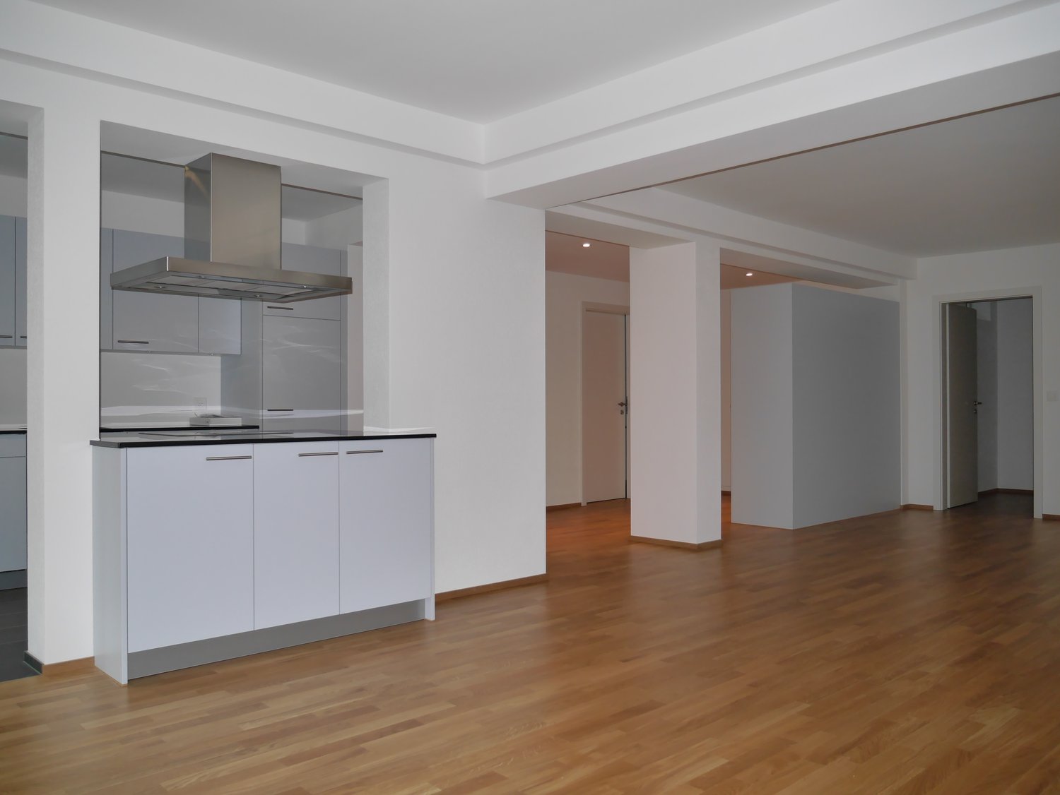Empty room, white walls, wooden floor, modern kitchen area with stainless steel extractor hood, white cabinets, black countertops