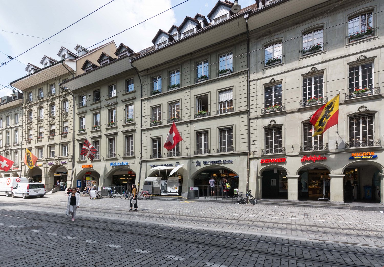 City street, multi-story building, flags on the top, street tram, pedestrians, bicycles, storefronts, white vans parked on the side