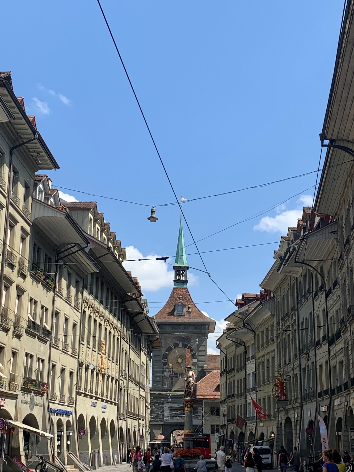 Swiss street, multiple story buildings, green spire, tower with clock, people walking