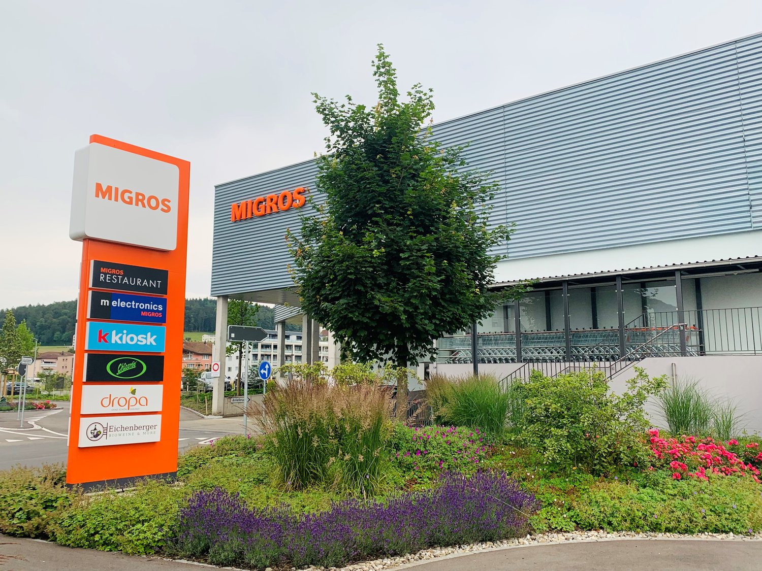 Large Migros supermarket building with several service stores, tree, and well-maintained flower beds in front