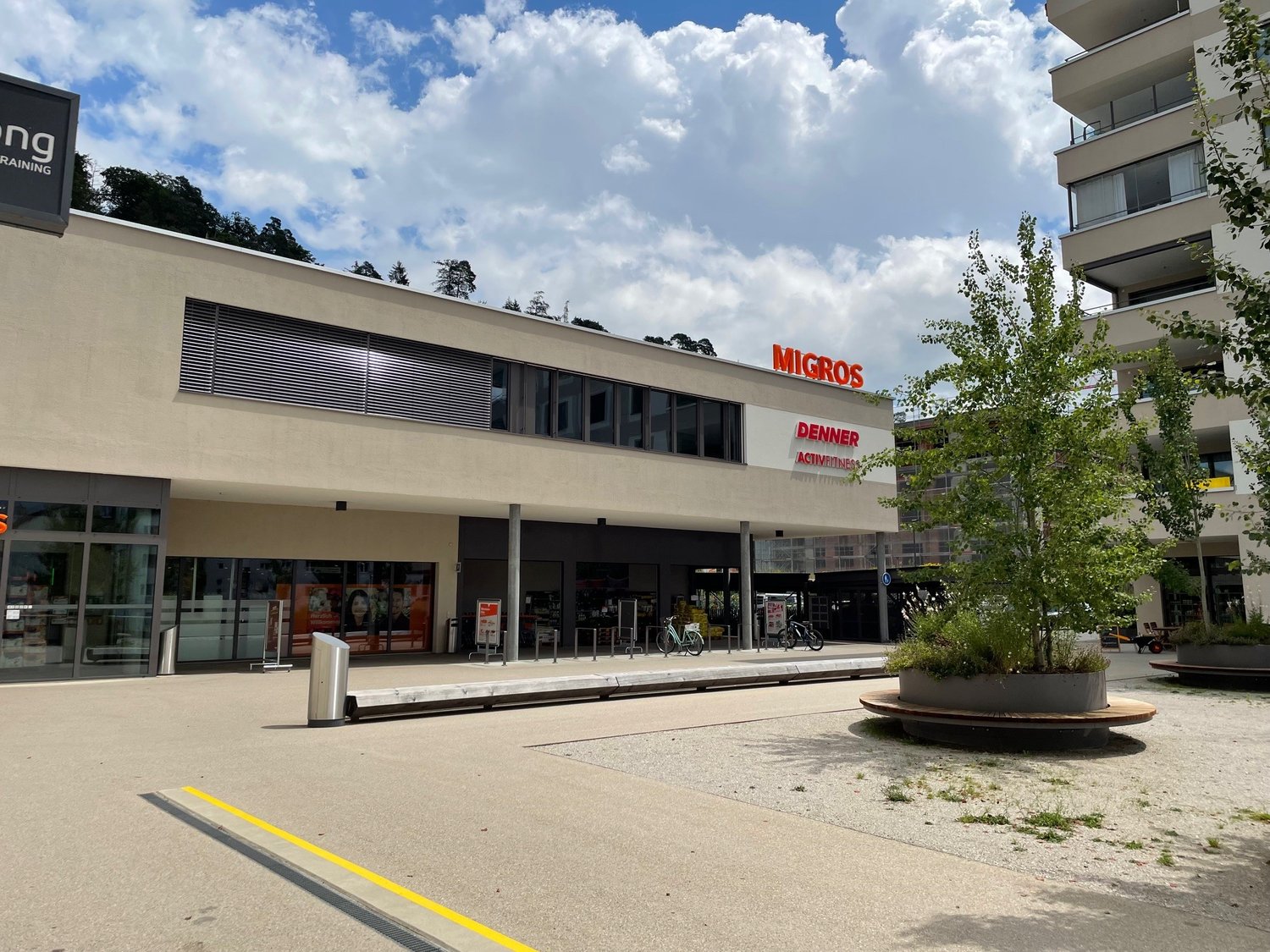 The building features a modern design with large glass windows and metallic pillars. It has a large entrance with automatic doors, a metal bollard barrier, and a bike rack in front. The entrance has a sign that reads 'Migros', indicating it might be a sup