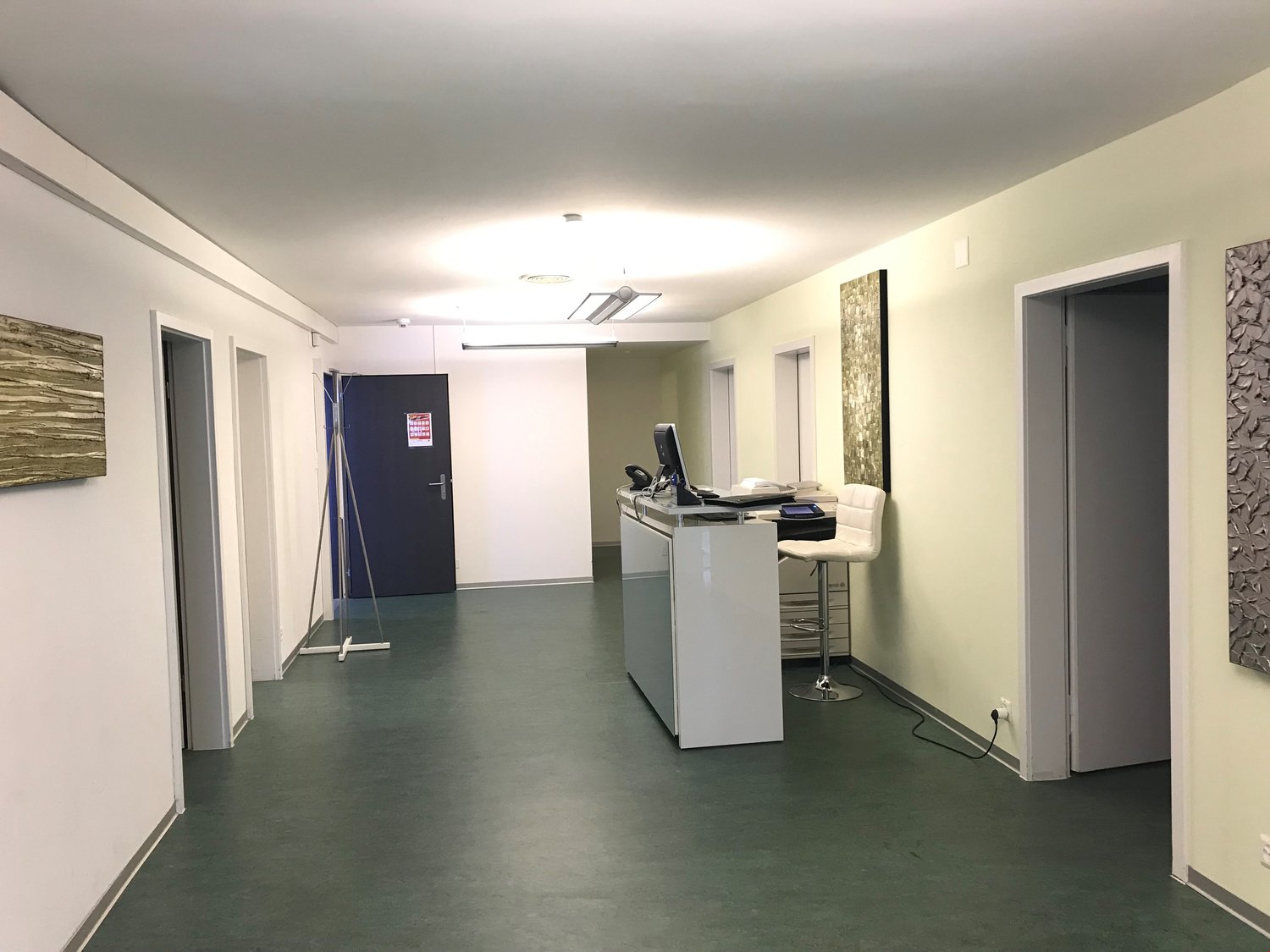 The image shows an office space with a reception desk, computer equipment, and a dark blue door. The walls are light-colored, and the floor is a gray concrete-like material. The space appears to be well-lit and organized.