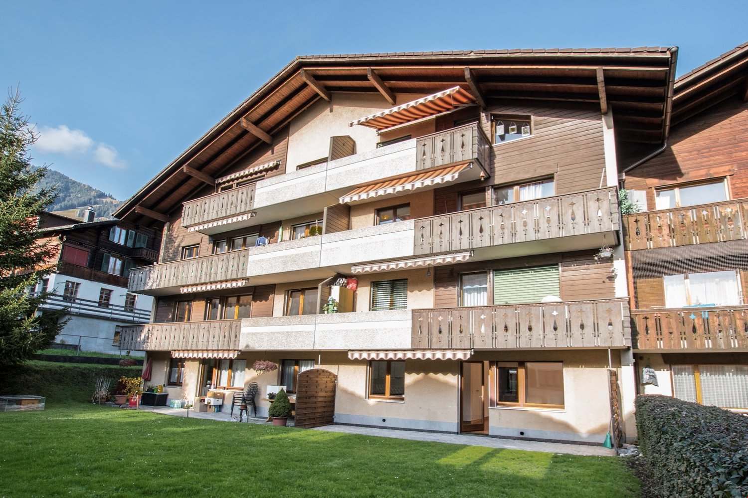 3-story apartment building with wooden and stone exterior, balconies on each floor, surrounded by a grassy lawn