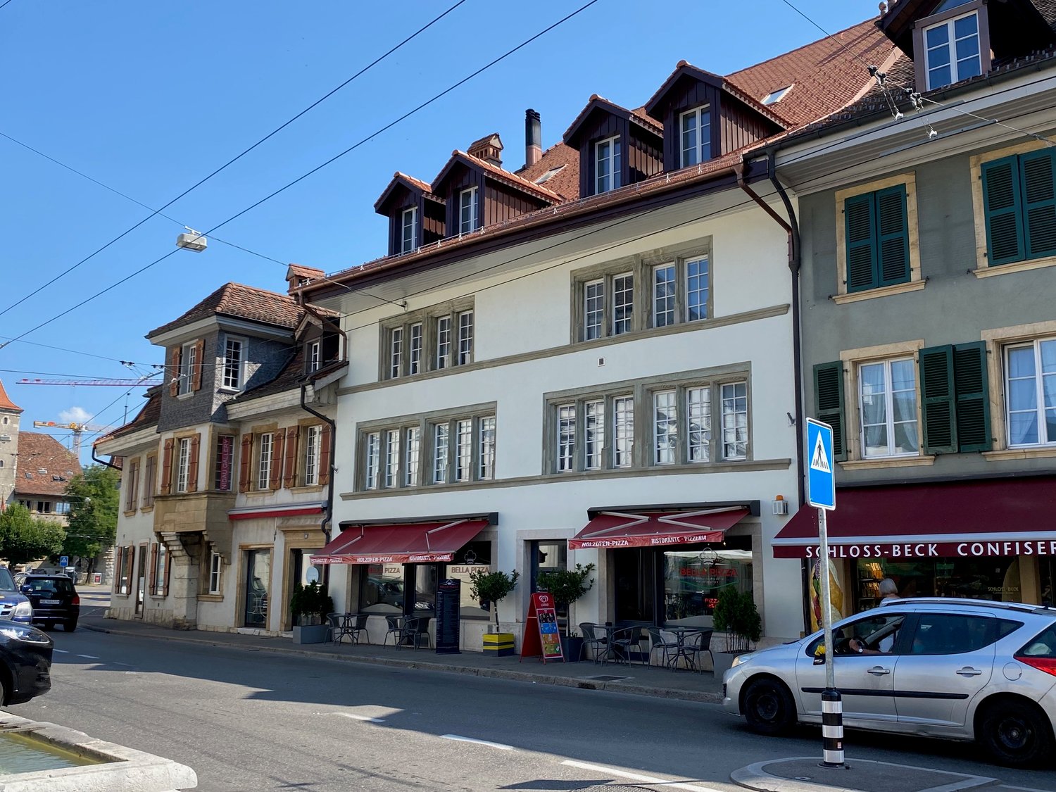 row of buildings, some with awnings, street with parked cars, greenery, and electric cables overhead