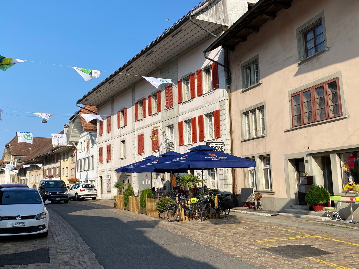 This image shows a street view of a quaint European town. The buildings have traditional architecture with red shutters and wooden beams. There are several restaurants and shops along the street, with outdoor seating areas under blue umbrellas. Bicycles a