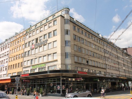 A multi-story commercial building with a distinctive architectural style, featuring rows of windows and a prominent corner entrance. The building appears to be located in an urban setting with other buildings and pedestrians visible in the surrounding are