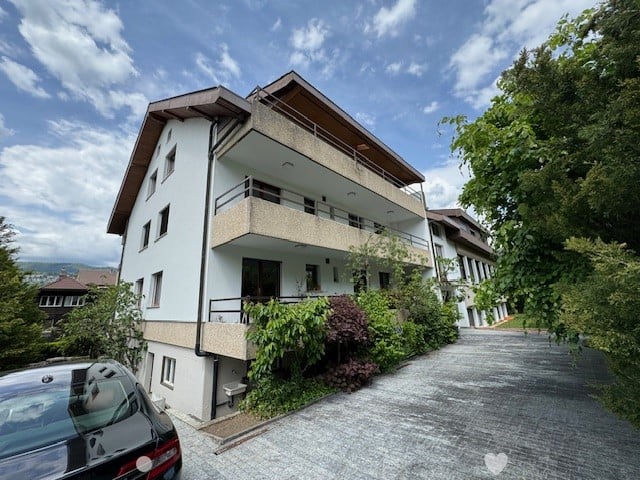 3-story apartment building with balconies, surrounded by trees and greenery, with a paved walkway leading to the entrance.