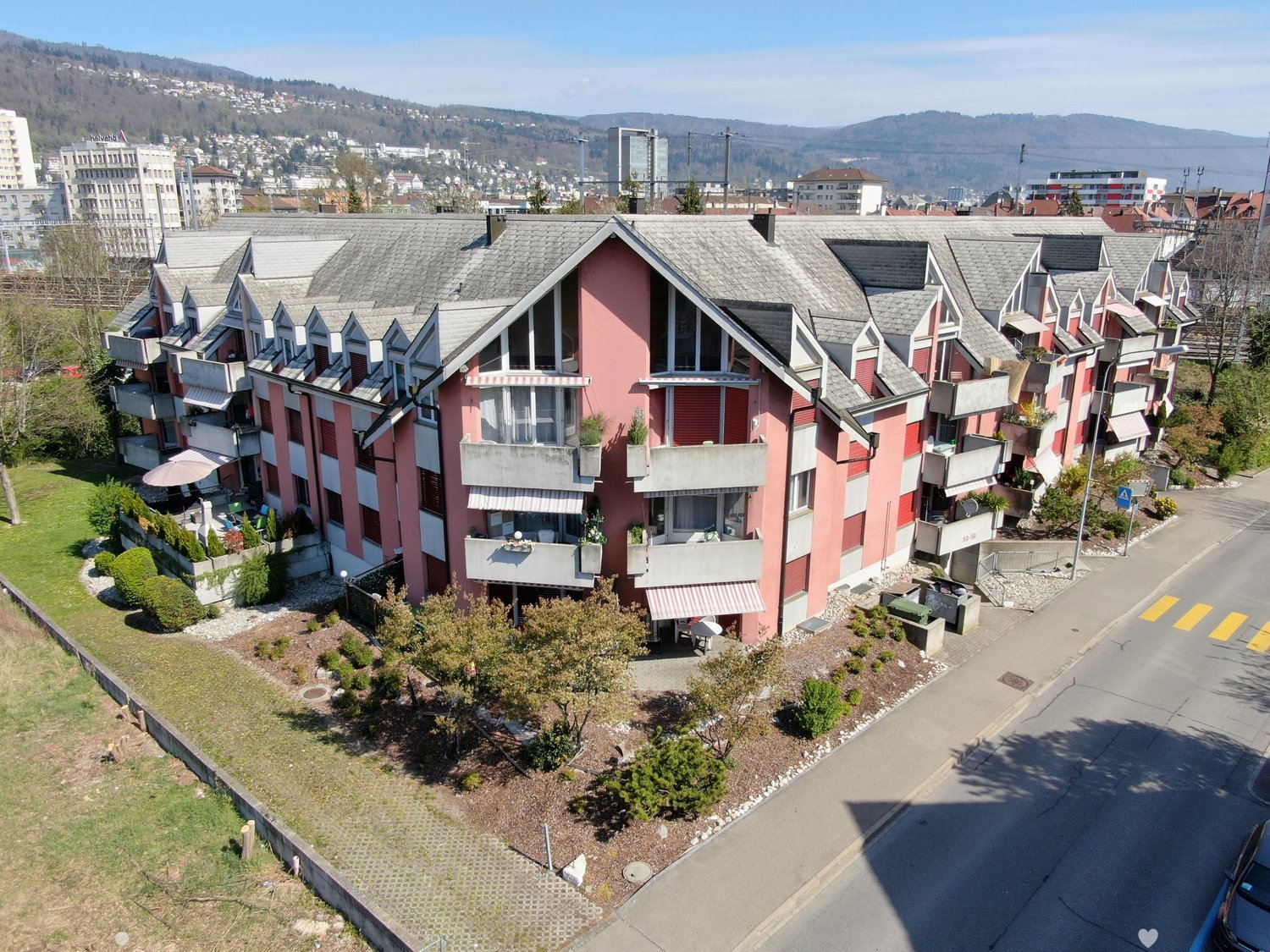 Multi-story apartment building with red and white exterior, pitched roofs, balconies, and landscaped grounds. The building is situated on a hillside overlooking a city skyline.