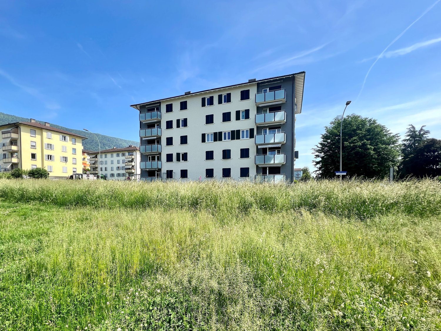 Multi-story apartment building with balconies, surrounded by a grassy field with trees in the background