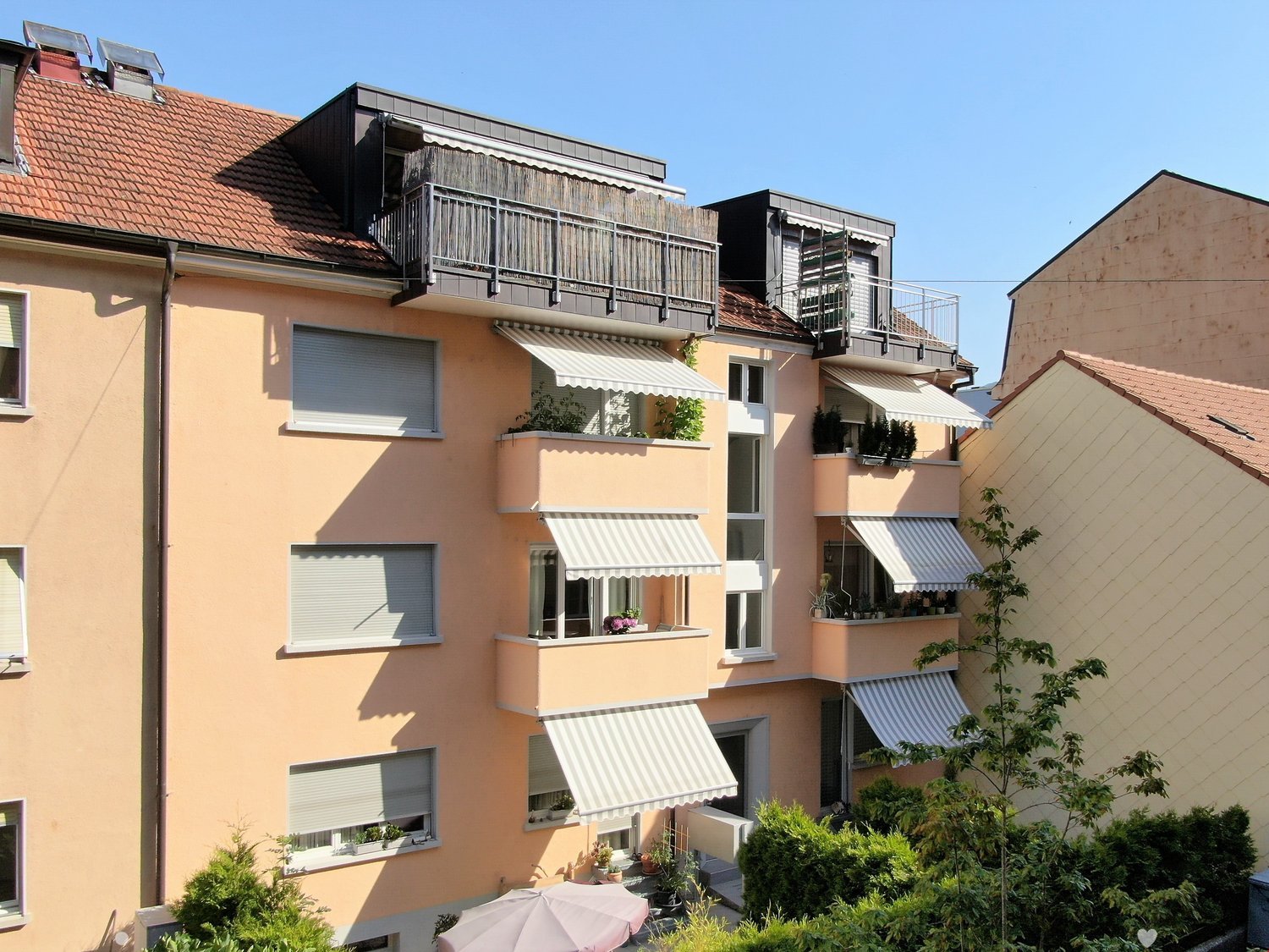 Three story beige building with tiled red roof, many balconies with white awnings, plants on the balconies