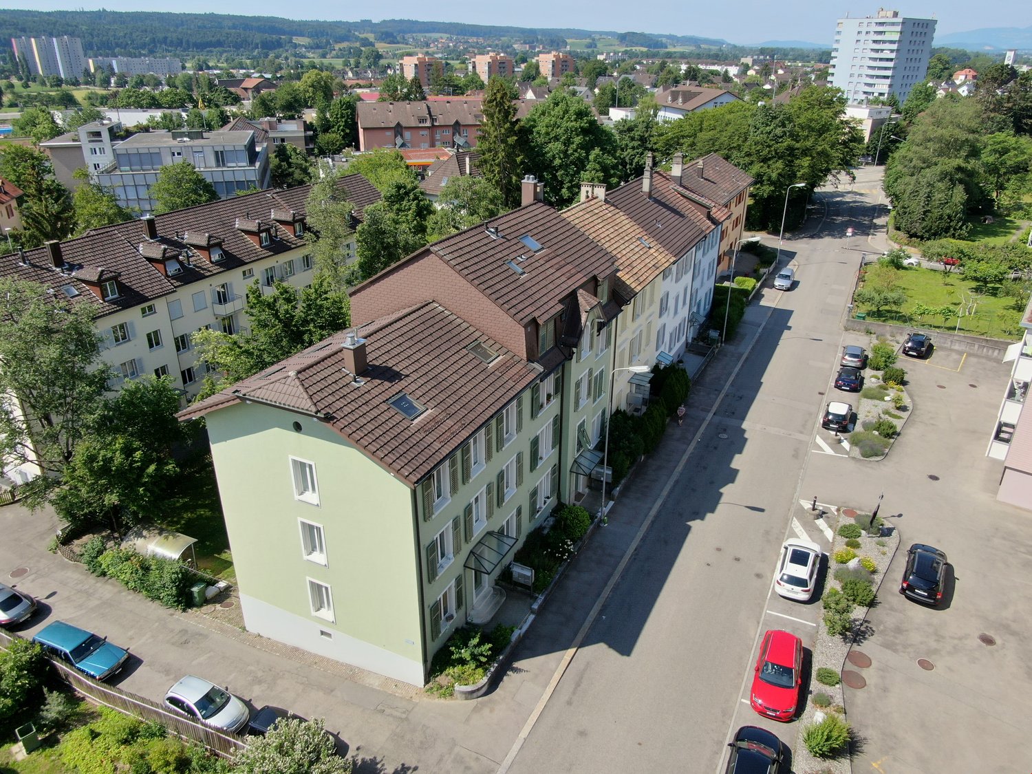 Aerial view of multiple residential buildings with brown roofs and white walls, surrounded by trees and greenery. Several cars parked in front of the buildings. A row of houses with a wide driveway and a street with cars driving and parked.