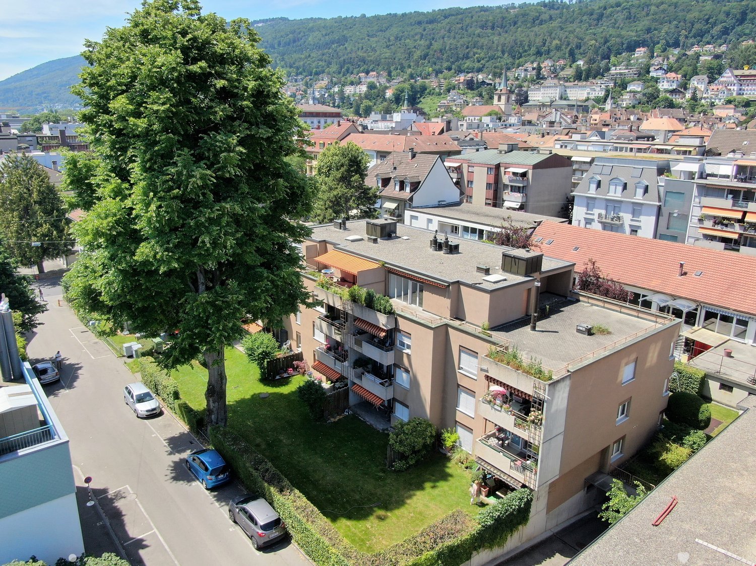 apartment building, multiple balconies, green area, road, multiple trees, mountains in the background