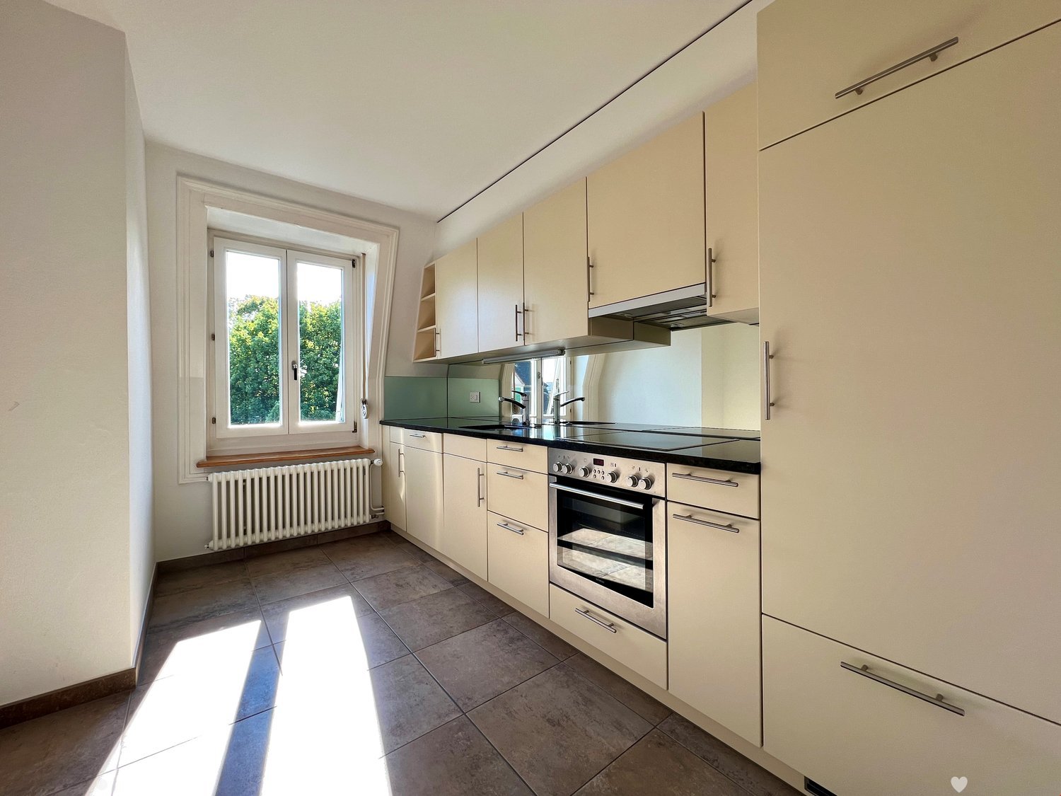 Spacious kitchen with beige cabinets, stainless steel appliances including a dishwasher, and tile flooring. Large window providing natural light.