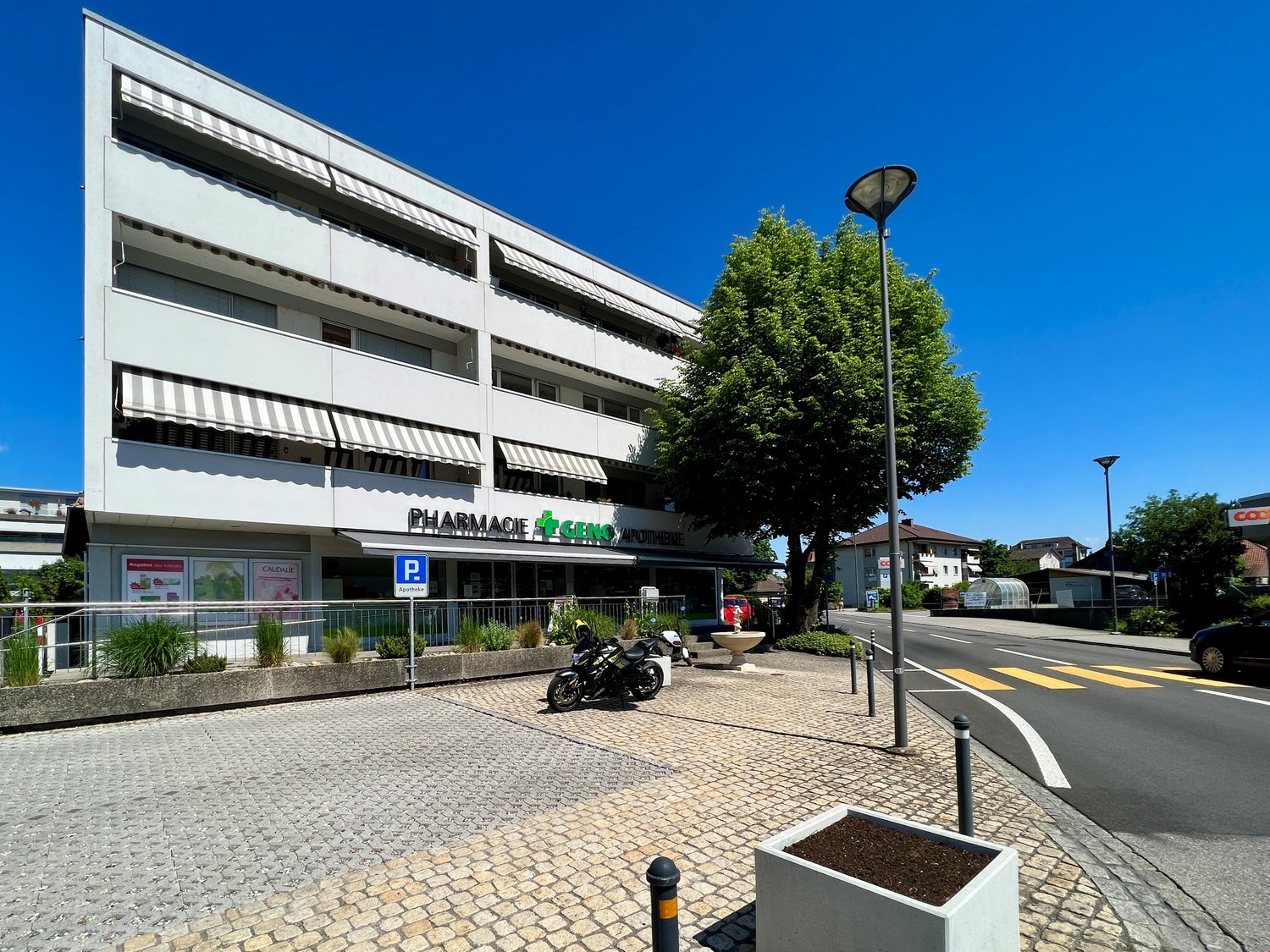 Apartment building, white exterior, several windows, awnings, small parking area, pharmacy sign, motorcycle parked