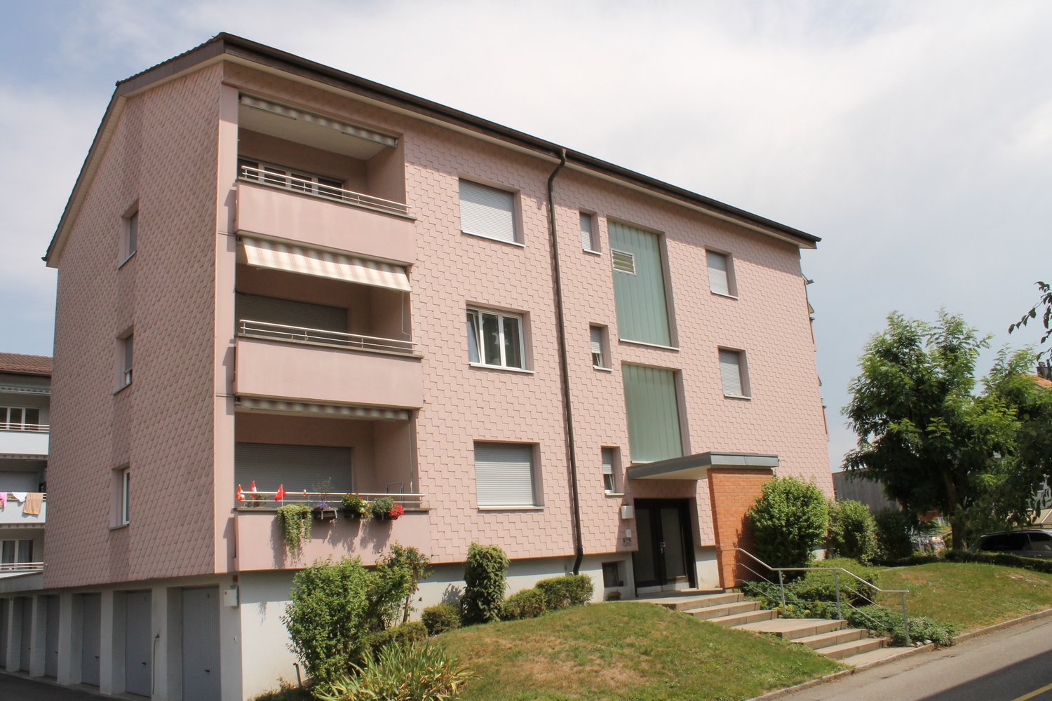 3-story apartment building with pink/peach colored exterior, balconies, large windows, and a landscaped yard in front.