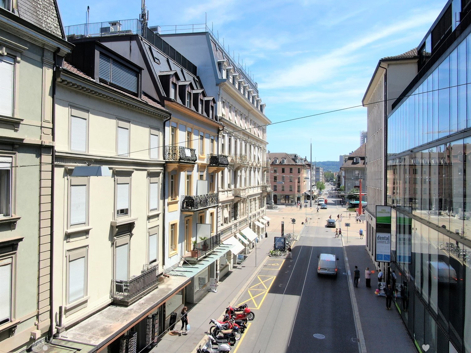 City street with multiple buildings on either side. Bicycles parked at side of road and scooters parked at sidewalk.