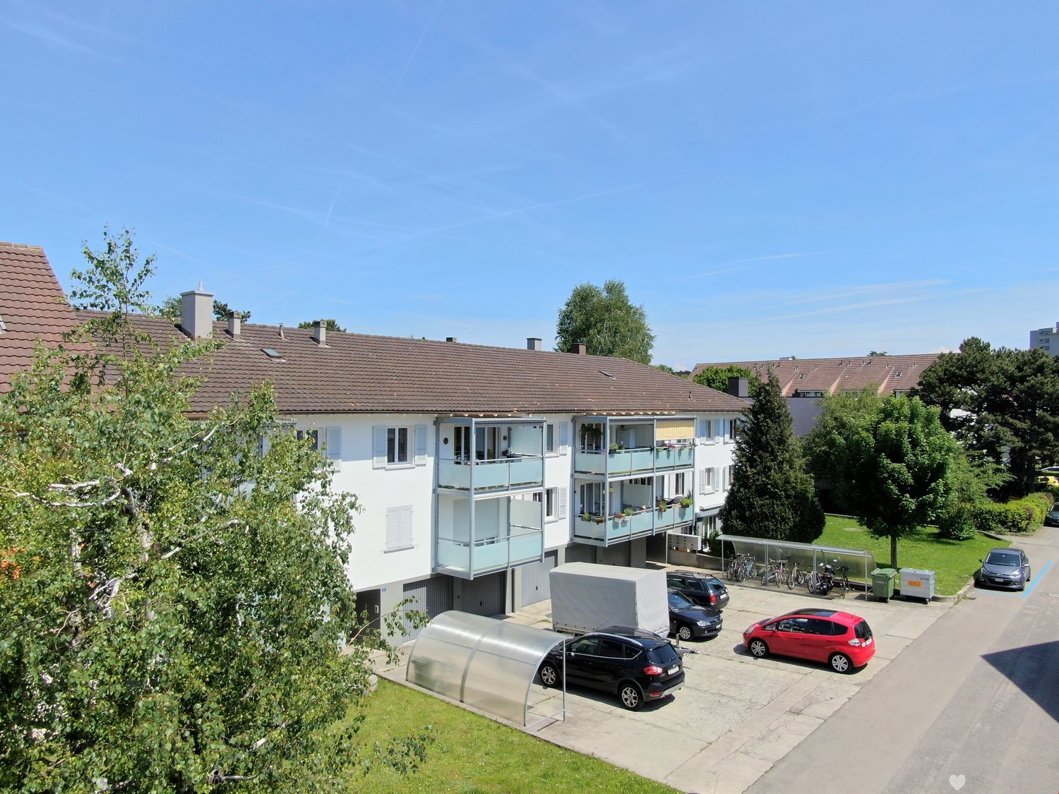Multi-story apartment building with balconies, surrounded by trees and greenery, with a parking area in the foreground