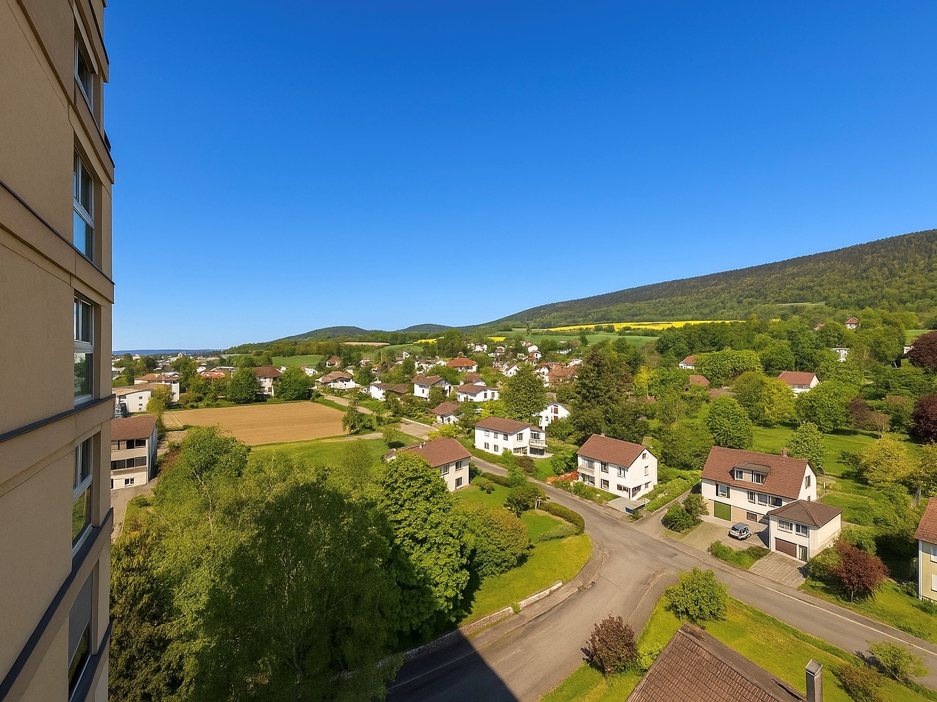 The image shows a scenic view of a small town or village nestled in a lush, green valley surrounded by rolling hills and mountains. The town is composed of residential houses and buildings with red roofs, surrounded by trees and greenery. In the distance,