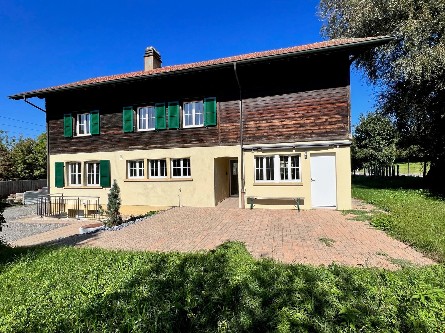 wooden and stone construction, terracotta roof, green shutters, brick driveway, bench, plants