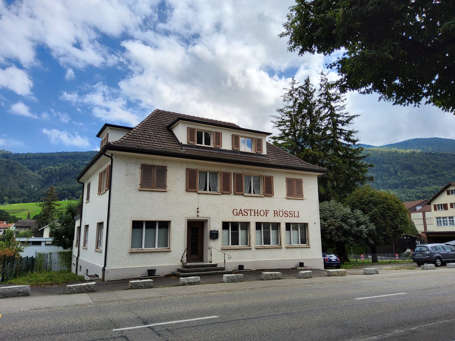 white house, brown roof, large windows, two cars parked, sign reads 'Gasthof Rossli', mountain in the background